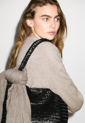 Sac tote en toile noir tissé avec une surface texturée et une sangle en tissu gris, exposé sur un fond clair. Modèle portant un pull beige.