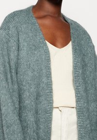 Lichtblauwe fuzzy cardigan met open voorkant en wijde mouwen. Gelaagd over een geribde crèmekleurige top, met zichtbare textuurcontrast.