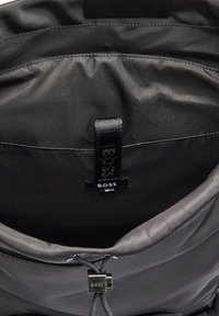 Black fabric bag interior with smooth texture, featuring a branded tag and an adjustable closure string. No visible compartments or patterns.