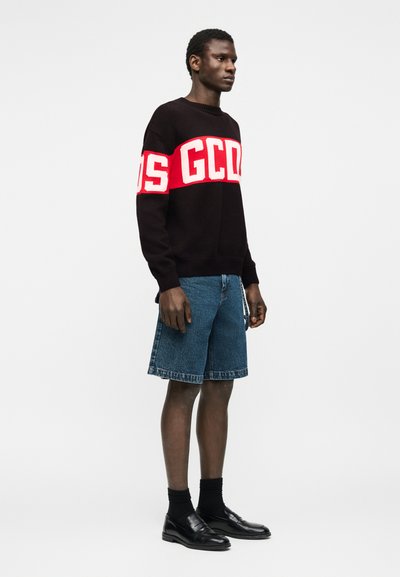 Άνδρας που φοράει μαύρο πουλόβερ με κόκκινα και λευκά γράμματα "GCDS", τζιν σορτς, μαύρες κάλτσες και μαύρα μοκασίνια, στέκεται μπροστά σε απλό φόντο.