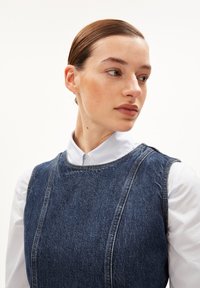 Salopette en denim sans manches, de couleur bleu profond, avec des coutures bien visibles et une texture usée, superposée sur une chemise blanche à col.