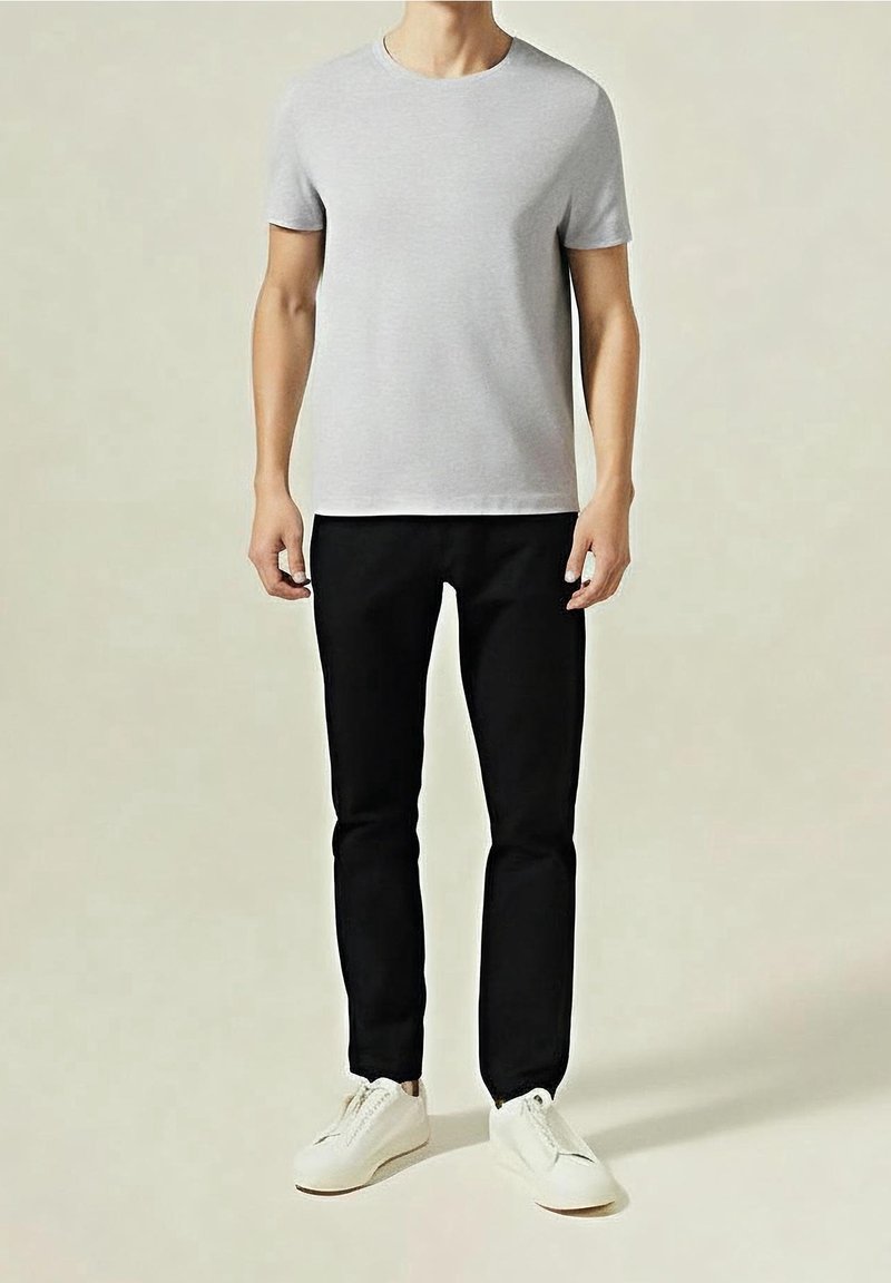 Homme debout portant un t-shirt gris clair à manches courtes, un pantalon noir et des baskets blanches sur un fond uni.