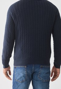 Marineblauer Strickpullover mit strukturiertem Zopfdesign, gerippten Bündchen und Saum, kombiniert mit klassischen blauen Jeans. Die Rückansicht zeigt die Details.