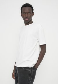 Άσπρο βαμβακερό μπλουζάκι με στρογγυλή λαιμόκοψη και κοντά μανίκια. Συνδυάζεται με σκούρα γκρι τζιν, με κλασικές λεπτομέρειες στις τσέπες και ραφές.