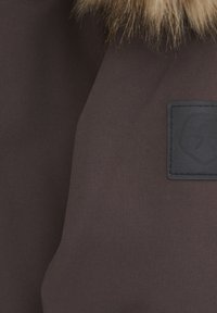 Brun jacka i texturerat tyg med en pälskrage och en svart lapp med en logotyp framtill.