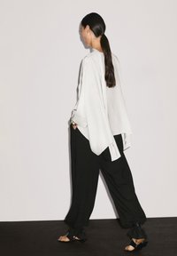 Blouse blanche surdimensionnée avec des manches fluides, associée à un pantalon noir ample avec des accents froncés aux chevilles et des sandales noires.