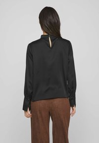 Schwarze Bluse mit hohem Kragen und einem Schlüsselhole-Detail hinten, lange Ärmel mit Knopfmanschetten, hergestellt aus glattem, weichem Stoff. Kombiniert mit braunen Hosen.