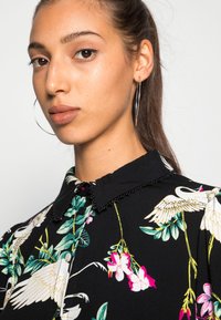 Camisa preta com colarinho, com um padrão floral que apresenta cisnes, folhagens e flores rosas; tecido texturizado com borda do colarinho desfiada.