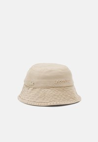 Beige bucket hat made of cotton. Features a textured surface and embroidered "LACOSTE" logo. The brim is soft and slightly wavy.