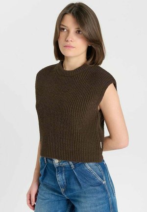Giovane donna con capelli corti castani che indossa un maglione a coste scuro senza maniche e jeans blu a vita alta, in piedi davanti a uno sfondo bianco.