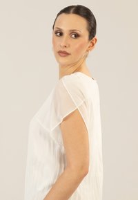Witte blouse met doorschijnende korte mouwen, geplooide textuur, ronde halslijn en knoopdetails op de rug. Soepele, lichte stof.