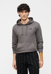 Šedá mikina s kapucňou s predným vreckom, sťahovacími šnúrkami a logom EA7. Nosená s čiernymi nohavicami s opakujúcim sa vzorom loga.