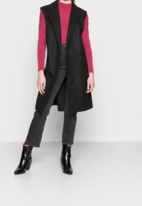 Black sleeveless coat with a structured design, paired with a fitted pink long-sleeve top and dark jeans. Black ankle boots complete the look.