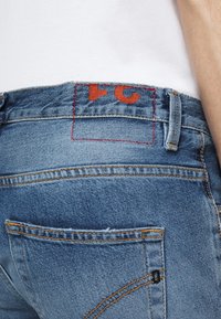Personne portant un jean bleu et une chemise blanche, gros plan sur la taille arrière avec une étiquette rouge cousue et des lettres partielles visibles au-dessus de la poche.