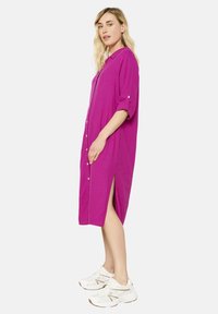 Abito fucsia con bottoni, maniche arrotolabili e spacchi laterali. Tessuto morbido e leggero con una vestibilità rilassata, abbinato a sneakers bianche.