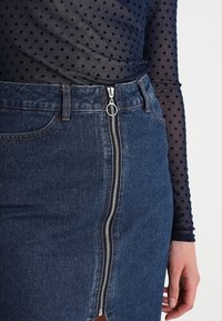 Gonna in denim con zip centrale in argento e accessori circolari, lavaggio medio e design aderente. Accoppiata con una blusa trasparente a pois.