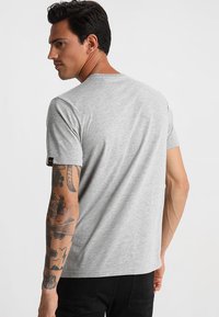 Camiseta gris de manga corta hecha de una tela suave, con cuello redondo y una pequeña etiqueta de la marca negra en la manga izquierda. Lleva puestos unos pantalones vaqueros negros.
