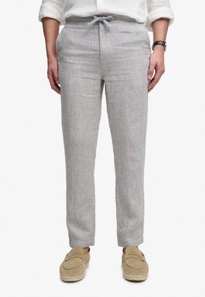 Osoba ubrana w jasno szare spodnie o prostych nogawkach z wiązanym pasem, beżowe mokasyny, białe podwinięte rękawy koszuli oraz czarny zegarek.