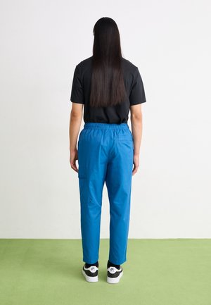 Person mit langen dunklen Haaren, die ein schwarzes T-Shirt, leuchtend blaue Hose und schwarze und weiße Nike-Sneaker trägt, steht auf grünem Boden vor einer weißen Wand.