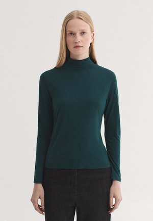 Frau mit glattem blondem Haar, die einen dunkelgrünen langärmeligen Rollkragenpullover und schwarze Hose vor einem schlichten hellen Hintergrund trägt.
