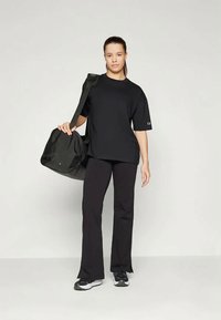 Fekete póló rövid ujjakkal, laza fazonban, fekete nadrággal és fekete sporttáskával párosítva. A táskát a vállán hordja.