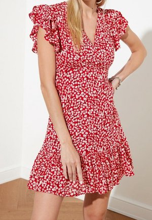 Vrouw draagt een korte rode jurk met witte madeliefjes, met vlindermouwen, een V-hals en een geruffelde zoom, staand binnen met hand op de heup.