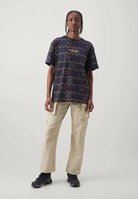 Trumpas rankovės tamsiai mėlynas marškinėliai su geometriniu raštu geltonos, raudonos ir mėlynos spalvos; derinami su smėlio spalvos kargo kelnėmis ir juodais sportbačiais.