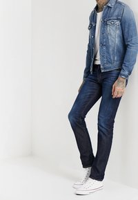 Jeansjacka i ljusblått, vit t-shirt under, mörkblå jeans och vita sneakers med stjärndetaljer; tatueringar synliga på armarna.