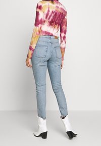 Jeans taille haute bleu clair avec une silhouette ajustée, présentant deux poches arrière. La tenue est associée à des bottes de cowboy blanches et un haut à manches longues tie-dye coloré.