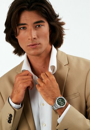 Silberne Armbanduhr mit grünem Ziffernblatt und silberner Hardware, getragen mit einem weißen Hemd und beigem Blazer. Der Stoff ist strukturiert und glatt.