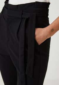 Detailní záběr na černé kalhoty s vysokým pasem a vázáním v pase; ruka odpočívající v kapse, ukazující malou tatérskou kresbu na zápěstí.