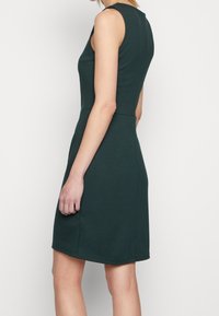 Bezramienna, ciemnozielona sukienka z dopasowanym gorsetem, lekko rozkloszowanym dołem i zaokrąglonym hemem; wykonana z gładkiego, elastycznego materiału.