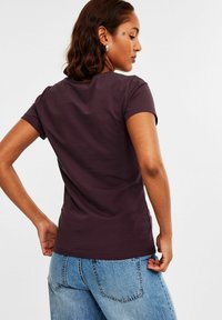 Kurzärmliges, figurschmeichelndes T-Shirt aus dunkelviolettem Baumwollstoff. Rundhalsausschnitt, glatte Textur, getragen mit hellblauen, eng anliegenden Jeans.