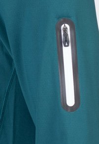 Chaqueta atlética en color teal que presenta un bolsillo con cremallera de color blanco y un tirador negro, fabricada con un tejido texturizado y transpirable.