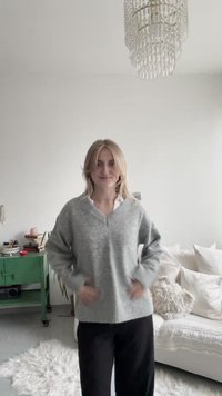 Pull gris clair à col en V avec texture côtelée et poches latérales, associé à un pantalon foncé, dans un intérieur lumineux et minimaliste.