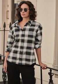 Černobílá kostkovaná košile s knoflíky a zakrčenými rukávy, doplněná černými kalhotami. Model má kulaté sluneční brýle a stříbrný prsten.