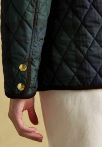 Jachetă matlasată de culoare verde închis cu un model în carouri, dotată cu butoane cu capse de aur și tivuri maro la manșete. Textură netedă și croială ajustată.