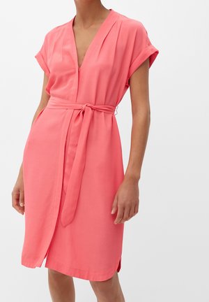 Žena v korálově růžových zavinovacích šatech po kolena s krátkými rukávy a zavázaným páskem v pase na neutrálním pozadí.