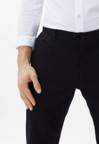 Pantaloni neri con una texture liscia, design aderente, tasche frontali e chiusura con bottone. Camicia bianca a maniche lunghe visibile.