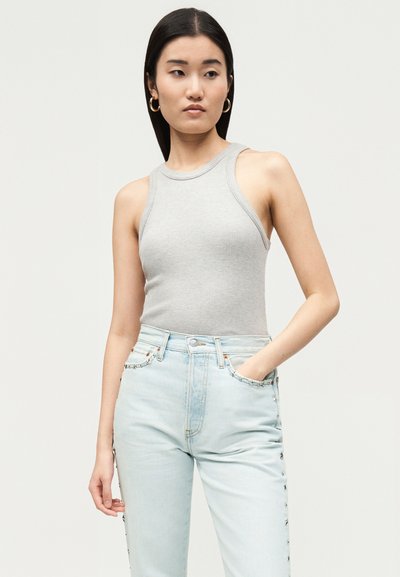 Femme aux longs cheveux foncés portant des boucles d'oreilles créoles dorées, un haut côtelé sans manches gris clair et un jean bleu clair, main dans la poche.