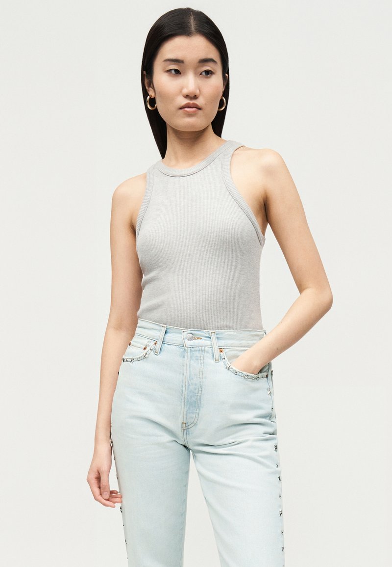 Femme aux longs cheveux foncés portant des boucles d'oreilles créoles dorées, un haut côtelé sans manches gris clair et un jean bleu clair, main dans la poche.