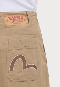 Pantalon beige avec une poche arrière présentant un motif brodé marron courbé et un patch en cuir de marque près de la taille.