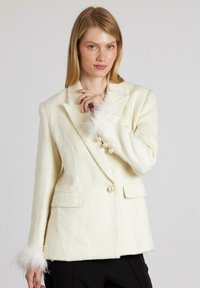 Blazer de marfil con una tela texturada, con puños de plumas, botones de perla y dos bolsillos frontales. Ajuste entallado con solapas de muesca.