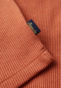 Tejido acanalado en un cálido color terracota con costuras visibles. Incluye una etiqueta azul con texto dorado. La textura parece suave y texturizada.