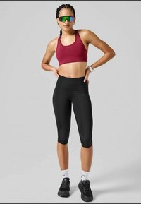 Athletic woman with braided hair in maroon sports bra, black capri leggings, white socks, black sneakers, and reflective sports sunglasses.