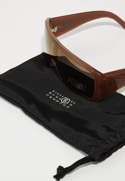 Lunettes de soleil de sport marron avec un design élégant, verres teintés dégradés et branches transparentes, reposant sur une pochette en tissu noir à motif numérique.