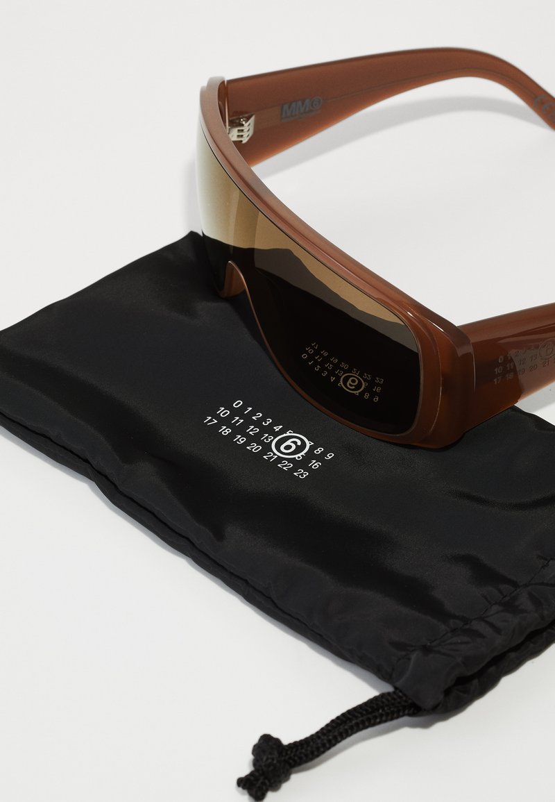 Lunettes de soleil de sport marron avec un design élégant, verres teintés dégradés et branches transparentes, reposant sur une pochette en tissu noir à motif numérique.