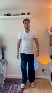 Fehér V nyakú póló és sötét, széles lábú nadrág szürke sportcipővel kombinálva. Belső tér színes világítással és egy polc, amelyen különböző tárgyak találhatók.