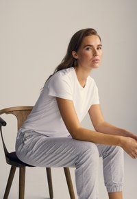 Hvid t-shirt med korte ærmer, parret med lysegrå joggerbukser med elastisk talje. Model siddende på en træstol med buet ryglæn.