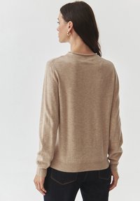 Beige gebreide trui met lange mouwen en een ronde hals, met ribgebreide manchetten en zoom. Het materiaal lijkt zacht en glad van textuur.